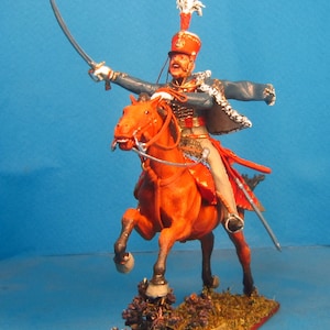 May include: A hand-painted miniature figure of a soldier on horseback, wearing a blue and red uniform with a white plume on his hat. The horse is a reddish-brown color and is in a rearing position. The figure is set on a base with brown and green foliage.