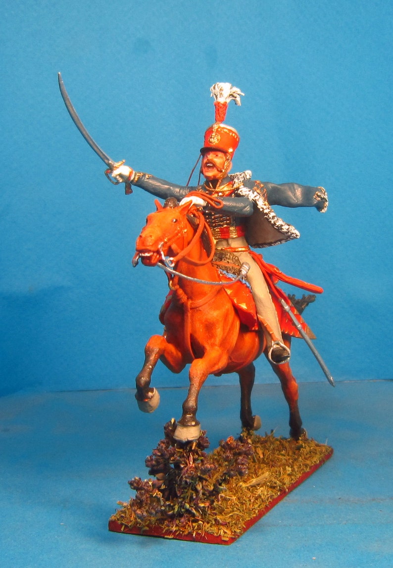 May include: A hand-painted miniature figure of a soldier on horseback, wearing a blue and red uniform with a white plume on his hat. The horse is a reddish-brown color and is in a rearing position. The figure is set on a base with brown and green foliage.