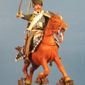 May include: A hand-painted miniature figure of a soldier on horseback, wearing a black and gold uniform with a white sash and a black bicorne hat with a red plume. The horse is brown and is in a rearing pose. The figure is set on a green base with brown dirt and purple flowers.