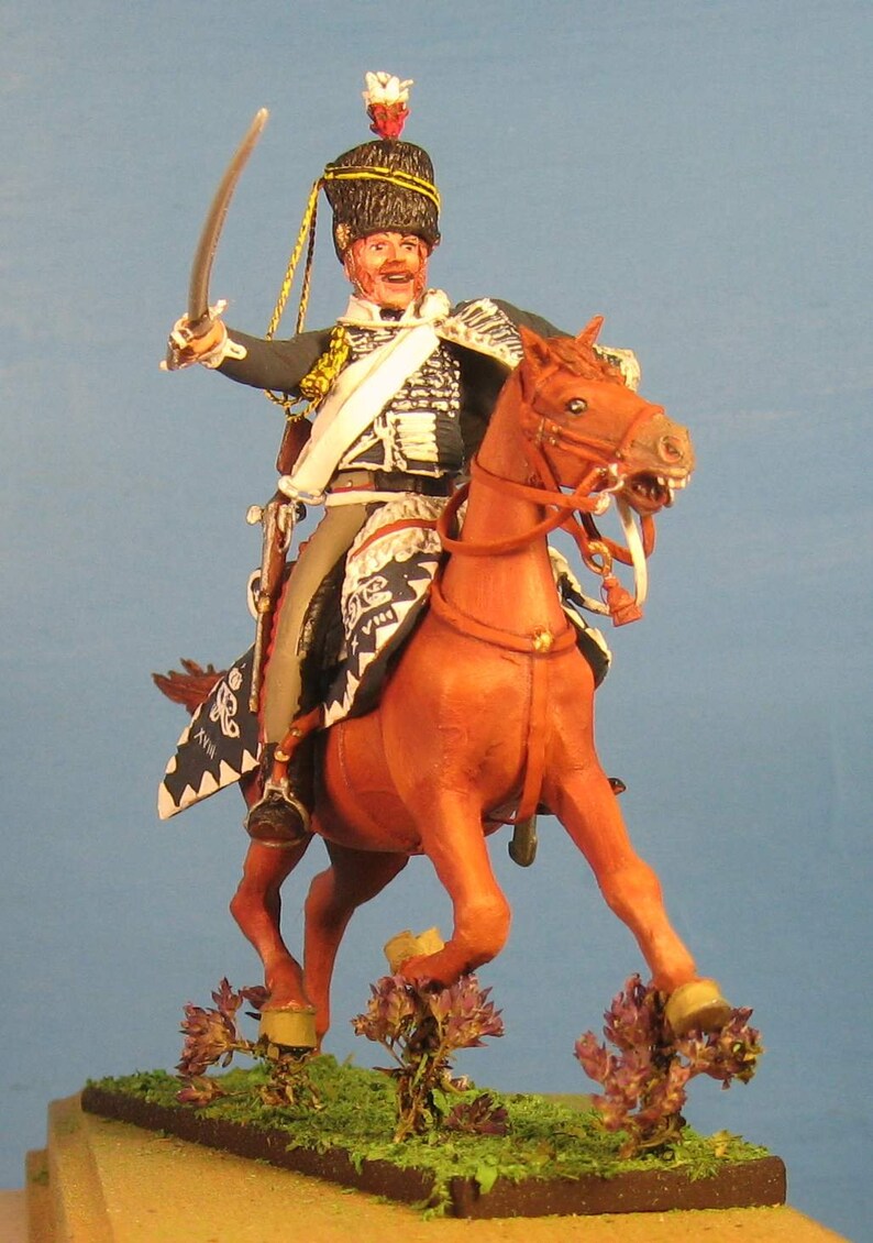 May include: A hand-painted miniature figure of a soldier on horseback, wearing a black and gold uniform with a white sash and a black bicorne hat with a red plume. The horse is brown and is in a rearing pose. The figure is set on a green base with brown dirt and purple flowers.