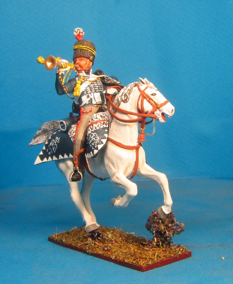May include: A hand-painted miniature figurine of a mounted soldier in a black and white uniform playing a trumpet. The soldier is riding a white horse with red and black saddle decorations. The miniature is set on a brown base.