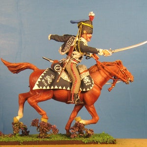May include: A hand-painted miniature figure of a cavalry soldier riding a brown horse. The soldier is wearing a blue and black uniform with a white sash and a black hat with a red plume. The horse is wearing a brown saddle and bridle. The figure is set on a green base with brown grass and small purple flowers.