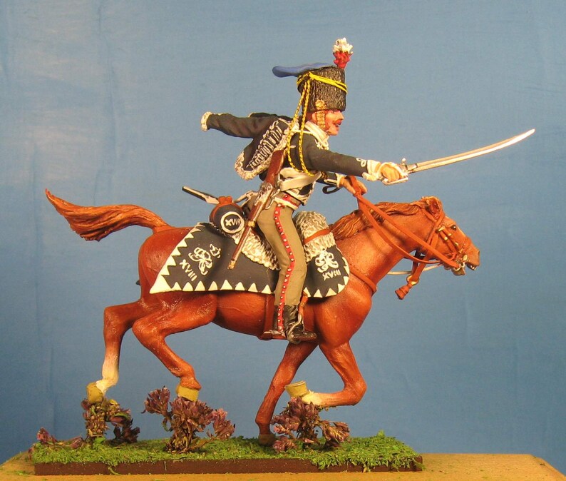 May include: A hand-painted miniature figure of a cavalry soldier riding a brown horse. The soldier is wearing a blue and black uniform with a white sash and a black hat with a red plume. The horse is wearing a brown saddle and bridle. The figure is set on a green base with brown grass and small purple flowers.