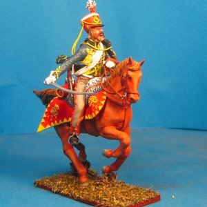 May include: A painted miniature figurine of a soldier on horseback, wearing a red and gold uniform with a tall, plumed hat. The horse is brown and has a red saddle. The figurine is posed in a dynamic action pose, with the soldier holding a sword.