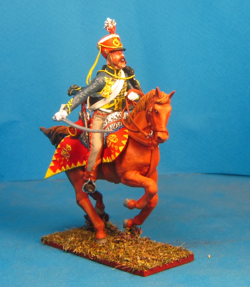 May include: A painted miniature figurine of a soldier on horseback, wearing a red and gold uniform with a tall, plumed hat. The horse is brown and has a red saddle. The figurine is posed in a dynamic action pose, with the soldier holding a sword.