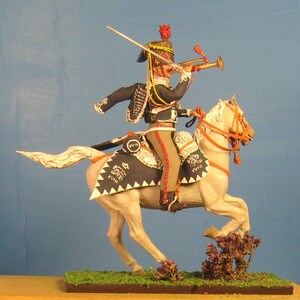 May include: A hand-painted miniature figure of a mounted soldier in a black and white uniform, holding a trumpet and riding a white horse with a black and white saddle. The soldier is wearing a red and gold hat with a plume. The horse is in a galloping pose.