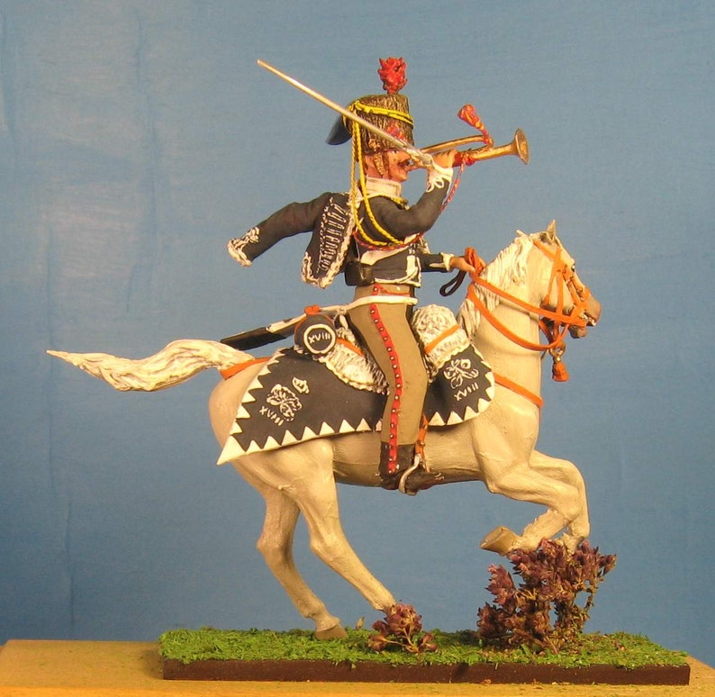 May include: A hand-painted miniature figure of a mounted soldier in a black and white uniform, holding a trumpet and riding a white horse with a black and white saddle. The soldier is wearing a red and gold hat with a plume. The horse is in a galloping pose.
