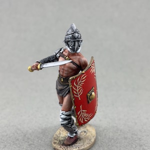 May include: A miniature gladiator figurine, clad in a silver helmet and armor, holds a sword and a red shield with gold accents. The figurine is posed in a fighting stance, showcasing intricate details of the armor and the gladiator's physique. The shield features a gold emblem.