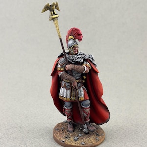 May include: A miniature Roman Centurion figure, featuring a red cloak, silver armour, and a plumed helmet. The figure holds a staff with a golden eagle at the top. The base is textured brown. The overall colour scheme is rich and historically accurate.
