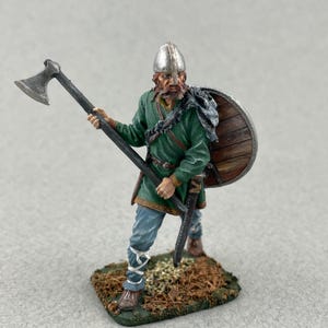 May include: A miniature Viking warrior figurine, holding an axe and shield. The figure wears a green tunic, blue pants, and a silver helmet. The shield is brown with a wooden texture. The base is a small patch of faux grass.