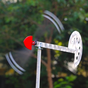 May include: A metal wind spinner mounted on a vertical pole, featuring a horizontal arm with a red semi-circular blade on one end and a stationary circular disc with perforated holes on the other, capturing motion blur from the spinning blade.