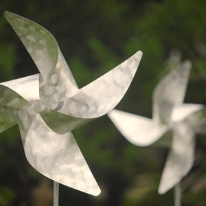 May include: Two silver pinwheels with four blades each, one in front of the other, against a blurred green background.