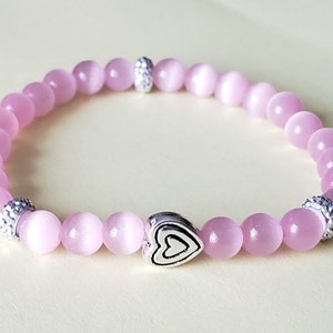 Puede incluir: Un brazalete de piedras preciosas rosas con un dije de corazón plateado y acentos de cuentas plateadas.