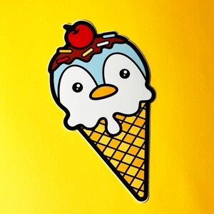Puede incluir: Un cono de helado de pingüino de dibujos animados con una cereza roja en la parte superior. El helado es azul y blanco con chispas de chocolate. El cono es amarillo y marrón.