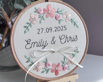 Peut inclure: Broderie sur tambour avec les noms "Emily & Chris" et la date "27.09.2025". Le motif comprend des fleurs roses et rose clair avec des feuilles vertes, et un nœud en ruban blanc. Le tambour est présenté sur un petit chevalet en bois.
