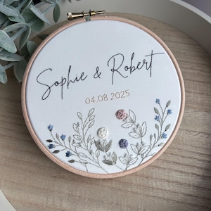 May include: Embroidered hoop art with the names "Sophie & Robert" and the date 04.08.2025 in black script. The white fabric is adorned with embroidered floral details in blue, pink, and white, set within a wooden hoop.