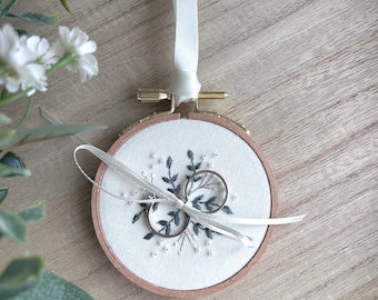 Peut inclure: Un tambour à broder circulaire en bois contenant un tissu blanc avec une broderie de feuilles vert foncé et de petites perles blanches. Deux anneaux en métal argenté sont fixés au centre du tissu par un fin ruban de satin blanc noué.