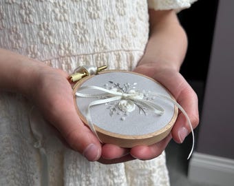 Peut inclure: Un tambour à broder circulaire en bois contenant un tissu gris clair avec des broderies florales blanches et de petites perles blanches. Un ruban de satin blanc est noué en boucle au centre du tissu, fixé par une vis de serrage en métal doré.