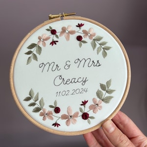 May include: A circular wooden embroidery hoop containing white fabric with a floral wreath design in shades of pink, burgundy, and green. The center features black embroidered text reading Mr & Mrs Creacy and the date 11.02.2024.