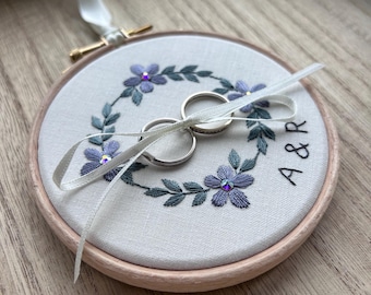 Peut inclure: Porte-anneaux brodé avec deux alliances en argent. Le support est un cercle avec un cadre en bois rose clair. Le tissu est brodé d'une couronne florale violette et verte, avec les initiales "A & R". Un ruban blanc est attaché.