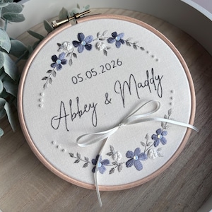 May include: Embroidered hoop art with the names "Abbey & Maddy" and the date "05.05.2026". The design features blue and white floral embroidery with a white ribbon bow. The hoop is set in a wooden tray.