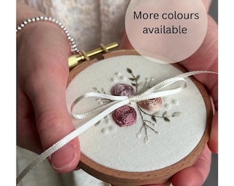 Peut inclure: Un tambour à broder circulaire en bois contenant un tissu blanc cassé avec des motifs floraux brodés à la main dans des tons rose poudré et mauve, agrémenté de petites perles blanches et d'un ruban de satin blanc noué. Texte : More colours available.