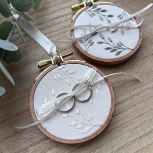 May include: Two circular wooden embroidery hoops containing white fabric with embroidered green and white leaf patterns. Each hoop features a white satin ribbon for hanging and a white ribbon tied in a bow, with two silver rings resting on one hoop.