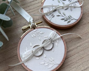 Peut inclure: Deux tambours à broder circulaires en bois contenant un tissu blanc avec des motifs de feuilles brodés en vert et blanc. Chaque tambour comporte un ruban de satin blanc pour suspendre et un ruban blanc noué, avec deux anneaux argentés.