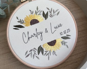 May include: Embroidered hoop art featuring sunflowers and foliage. The text "Charley & Lucie 12.12.25" is stitched in black script. The embroidery is set in a wooden hoop with a light pink inner ring, displayed on a wooden tray.
