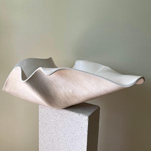 May include: A large, off-white ceramic bowl with a wide, undulating, and wavy rim. The exterior surface features a subtle, fine-lined texture, while the interior is smooth and matte. The bowl has a deep, organic, and asymmetrical shape.