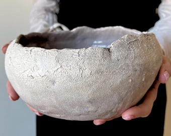 Puede incluir: Un cuenco de cerámica hecho a mano de forma redonda, con una superficie exterior gruesa, irregular y texturizada en tonos blanco roto o gris claro. El borde es irregular y dentado, mientras que el interior del cuenco parece liso con un acabado brillante.