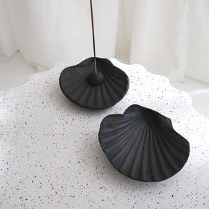 Könnte beinhalten: Zwei mattschwarze muschelförmige Räucherstäbchenhalter mit gerippter Textur. Ein Halter enthält eine kleine kugelförmige Perle, in die ein dünnes Räucherstäbchen vertikal eingesetzt ist. Die Objekte stehen auf einer weißen Oberfläche mit roten und schwarzen Sprenkeln.