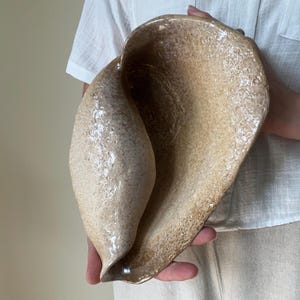 May include: A hand-built ceramic bowl with an organic, shell-like shape. The surface features a textured, speckled beige and tan glaze with a glossy finish. The form is asymmetrical, curving inward to create a deep, hollow interior space.
