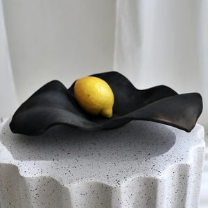 Peut inclure: Un bol noir de forme organique avec un citron jaune à l'intérieur. Le bol est posé sur un piédestal blanc texturé avec un motif ondulé. L'arrière-plan est un tissu blanc doux.