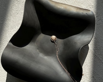 Può includere: Una ciotola in ceramica nero opaco con bordi ondulati e una base profonda e curva. Al centro poggia un piccolo medaglione circolare color oro metallizzato con un intricato motivo in rilievo, attaccato a una sottile catena a maglie dorata.