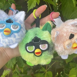 May include: Three plush Furby keychains with soft faux fur in light blue, lime green, and pale pink. Each features large plastic eyes with star or heart details, a yellow beak, and a plastic clip attached to a fabric loop for hanging.