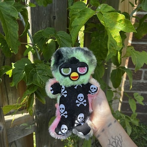 May include: A plush toy with long, shaggy fur in shades of black, mint green, and pink. The toy features a black plastic faceplate with large, round eyes containing green and pink star-patterned irises, and a yellow beak. It wears a black onesie with white skull and crossbones prints.