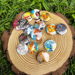 May include: A collection of circular pin-back badges featuring various colourful cartoon creature illustrations. The badges display diverse characters in shades of orange, blue, yellow, and grey, arranged in a cluster on a circular wooden slice.