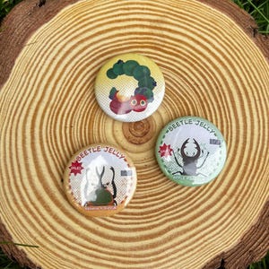 May include: Three circular pinback buttons. One features a green caterpillar and red fruit. The other two are labelled Beetle Jelly, one in tan with a rhinoceros beetle and banana flavour text, the other in green with a stag beetle and apple flavour text.
