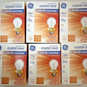 Puede incluir: Seis cajas de bombillas de bajo consumo GE Crystal Clear. Cada caja contiene dos bombillas y está etiquetada con la potencia, el brillo, el coste energético estimado y la vida útil. Las cajas son todas blancas con un fondo amarillo y naranja.