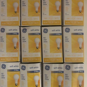 Op de afbeelding: Twaalf 3-weg-gloeilampen in zacht wit. De lampen zijn verpakt in doorzichtig plastic en hebben een geel etiket met het GE-logo en productinformatie. De lampen zijn geschikt voor 50/200/250 watt en hebben een levensduur van 1,1 jaar.