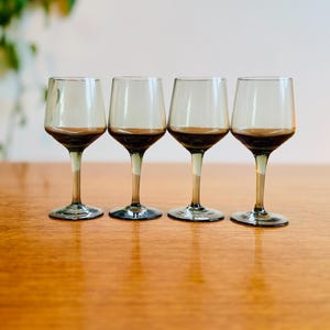 May include: Four clear glass wine glasses with a smoky grey tint. The glasses are arranged in a row on a wooden surface.