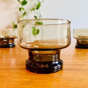 Op de afbeelding: Drie vintage bruinglazen drinkglazen met een voet. De glazen hebben een afgeronde komvorm en een donkerbruine kleur. De glazen staan op een houten oppervlak.