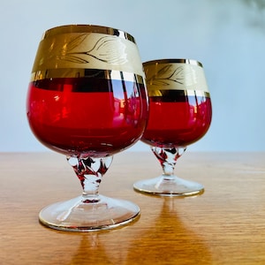 May include: Two red glass brandy snifters with gold trim and a white floral design. The glasses have a twisted stem and a round base.