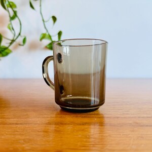 May include: A clear, brown-tinted glass mug with a handle and three small, dark brown decorative dots. The mug sits on a wooden surface, with green leafy vines in the background.