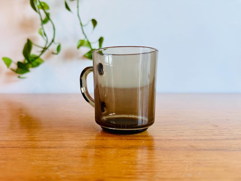 May include: A clear, brown-tinted glass mug with a handle and three small, dark brown decorative dots. The mug sits on a wooden surface, with green leafy vines in the background.