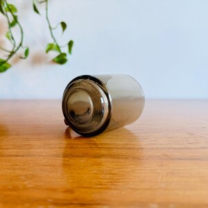 May include: A clear, cylindrical glass with a dark gray tint, lying on its side. The bottom of the glass is visible, showing a circular base. The glass is resting on a wooden surface.