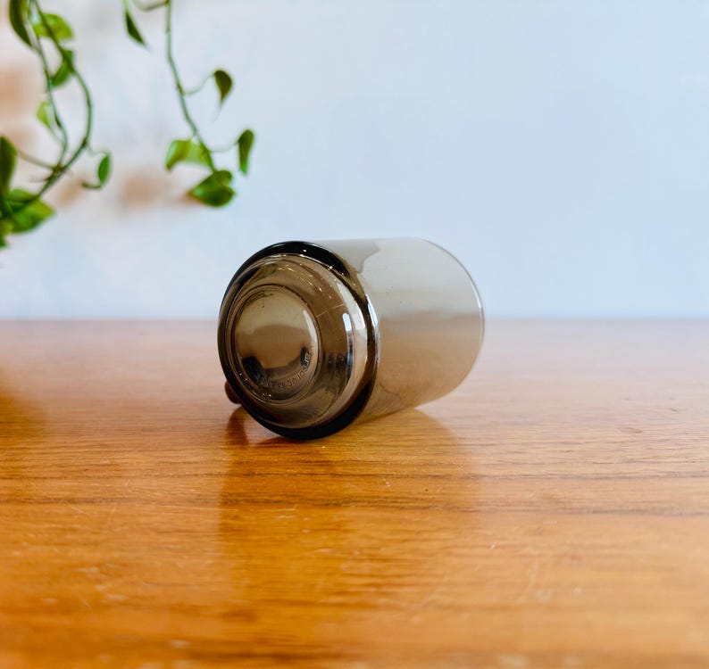 May include: A clear, cylindrical glass with a dark gray tint, lying on its side. The bottom of the glass is visible, showing a circular base. The glass is resting on a wooden surface.