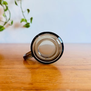 May include: A clear glass mug with a black rim and handle. The mug is sitting on a wooden surface. The mug is empty and appears to be clean. The mug is a vintage style.