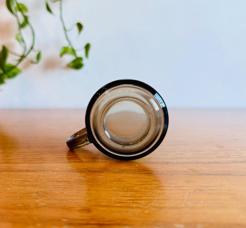 May include: A clear glass mug with a black rim and handle. The mug is sitting on a wooden surface. The mug is empty and appears to be clean. The mug is a vintage style.